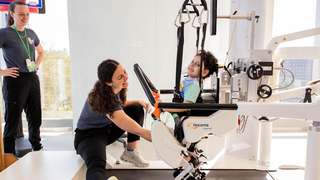A child using a machine trying to learn how to walk, assisted by two hospital staffs