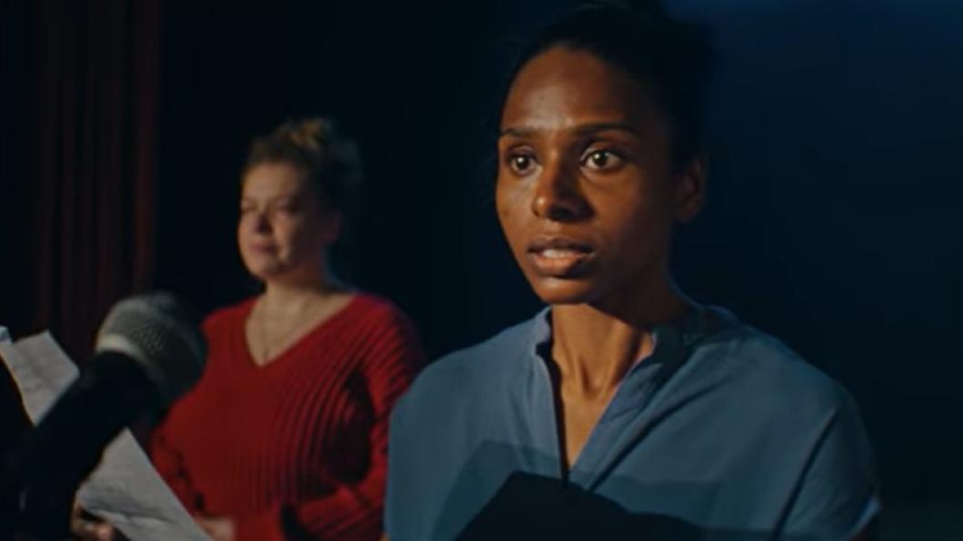 Woman in blue shirt stands in front of microphone while another woman is in the background