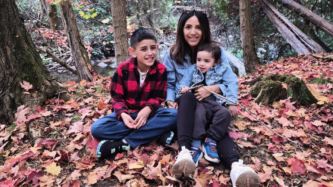 Two boys with dark hair sit in lap of woman with dark hair among colourful fall leaves