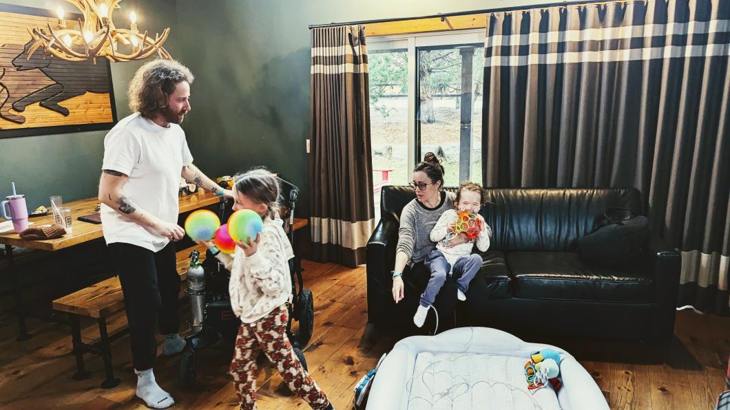 Woman holding child on couch while another girl walks with coloured balls in front of a man