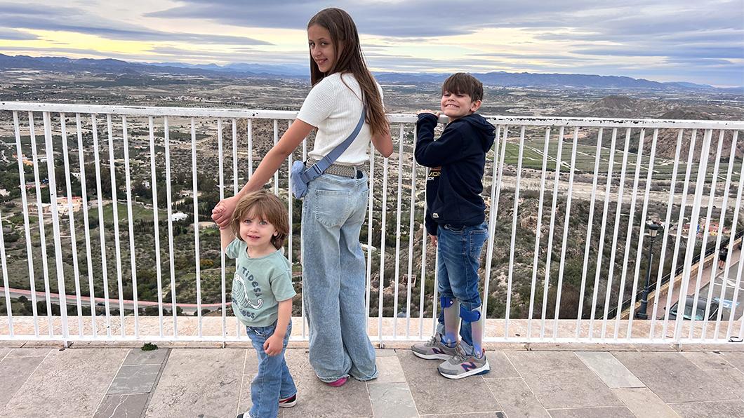 An older sister and her two younger brothers stand on a balcony.