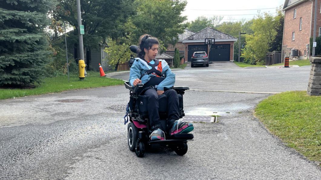 Teen with long dark hair drives power wheelchair in suburban neighbourhood.