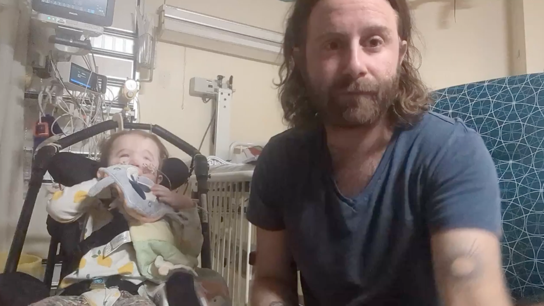 Man with beard beside child in stroller in hospital room with medical equipment