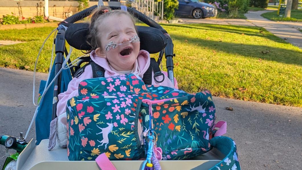 Young girl with oxygen prongs and huge smile sits in wheelchair with backpack on tray.
