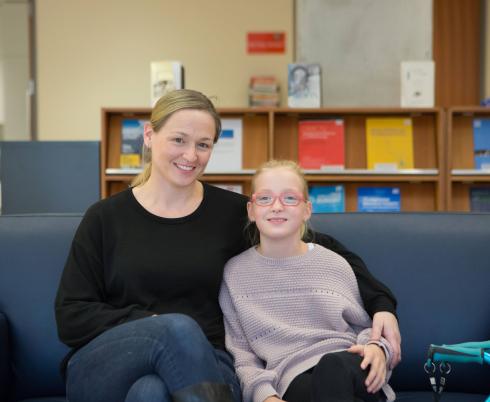A mother and daughter sit together on a blue couch. The mother has blonde hair and wears a black sweater and blue jeans. The daughter has blonde hair and wears a grey sweater and pink glasses. Her mobility device can be seen at the corner of the image.