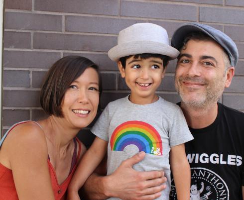 A woman with short brown hair is smiling and hugging her young son. The young boy is smiling and wearing a grey t-shirt with a rainbow on it. His father is kneeling beside him and holding him. He is wearing a black t-shirt and a baseball cap.