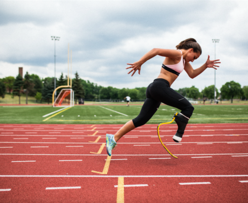 A young woman running on a track. She is wearing a running blade on her right leg, black leggings and a pink top.