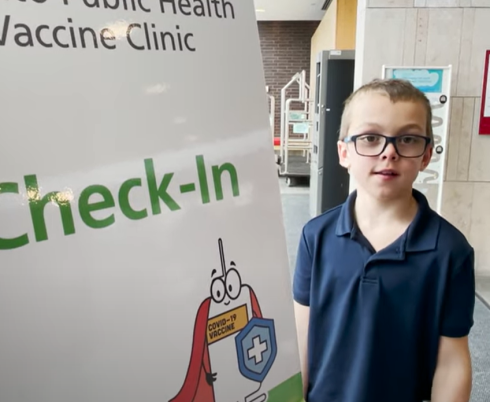 Client ambassador Alex Winter standing next to check-in sign for a vaccine clinic at Holland Bloorview's.