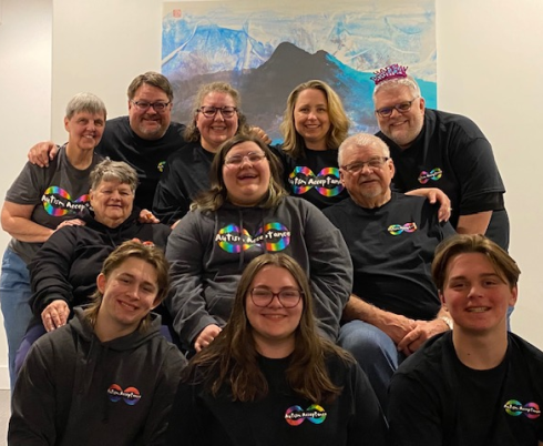 A smiling teen and their family sitting together for a family photo. Each family member is smiling and wearing black t-shirts and sweaters that say, "Autism Acceptance."