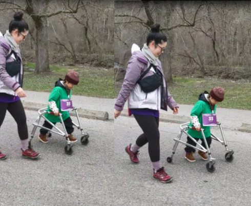 A young child walking on pavement. He is using a walker and is wearing a green t-shirt, brown shoes and a red beanie hat. His mom is walking beside him. She is wearing her hair in a bun, a purple and white rain jacket and black athletic pants.