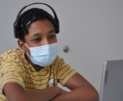 A boy with brown skin tone and black hair in a striped shirt, wearing a mask and headphones seated in front of a computer