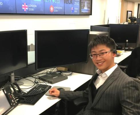 A young person sitting in front of a computer screen and smiling. They are wearing a black suit jacket with white stripes and black glasses.