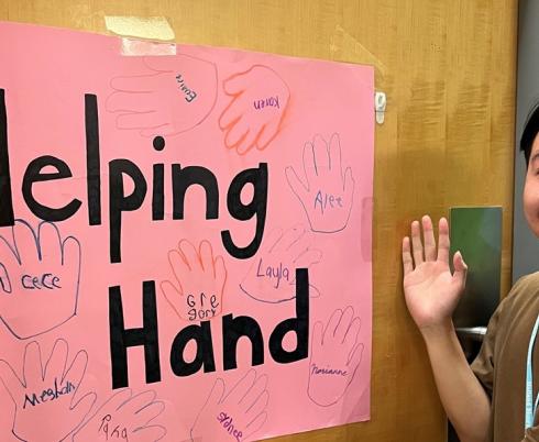 An adult volunteer holding the right hand in front of a hand draw poster with some hands on, and the words "Helping Hand"