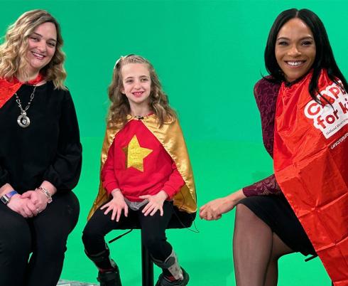 Two adults and a child with their red capes sitting in front of a green background for TV airing