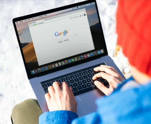 Person wearing a blue hoodie and orange toque is sitting with a grey laptop open and browsing on the Internet.