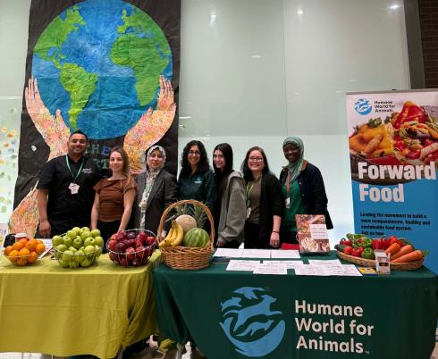 Photograph of seven people standing behind two tables displaying fresh fruits and vegetables at a Humane World for Animals event promoting sustainable, healthy, plant-based food systems. Tables feature green and yellow tablecloths, baskets of apples, bananas, and carrots, with a large poster and a backdrop of a painted Earth held by hands.