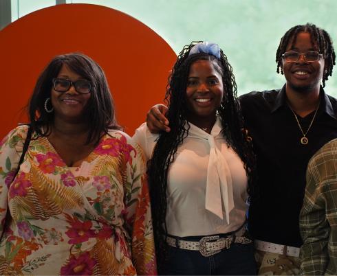 Photograph of four people standing side by side in front of a large orange circular backdrop with a green blurred background. They are wearing casual clothing, 